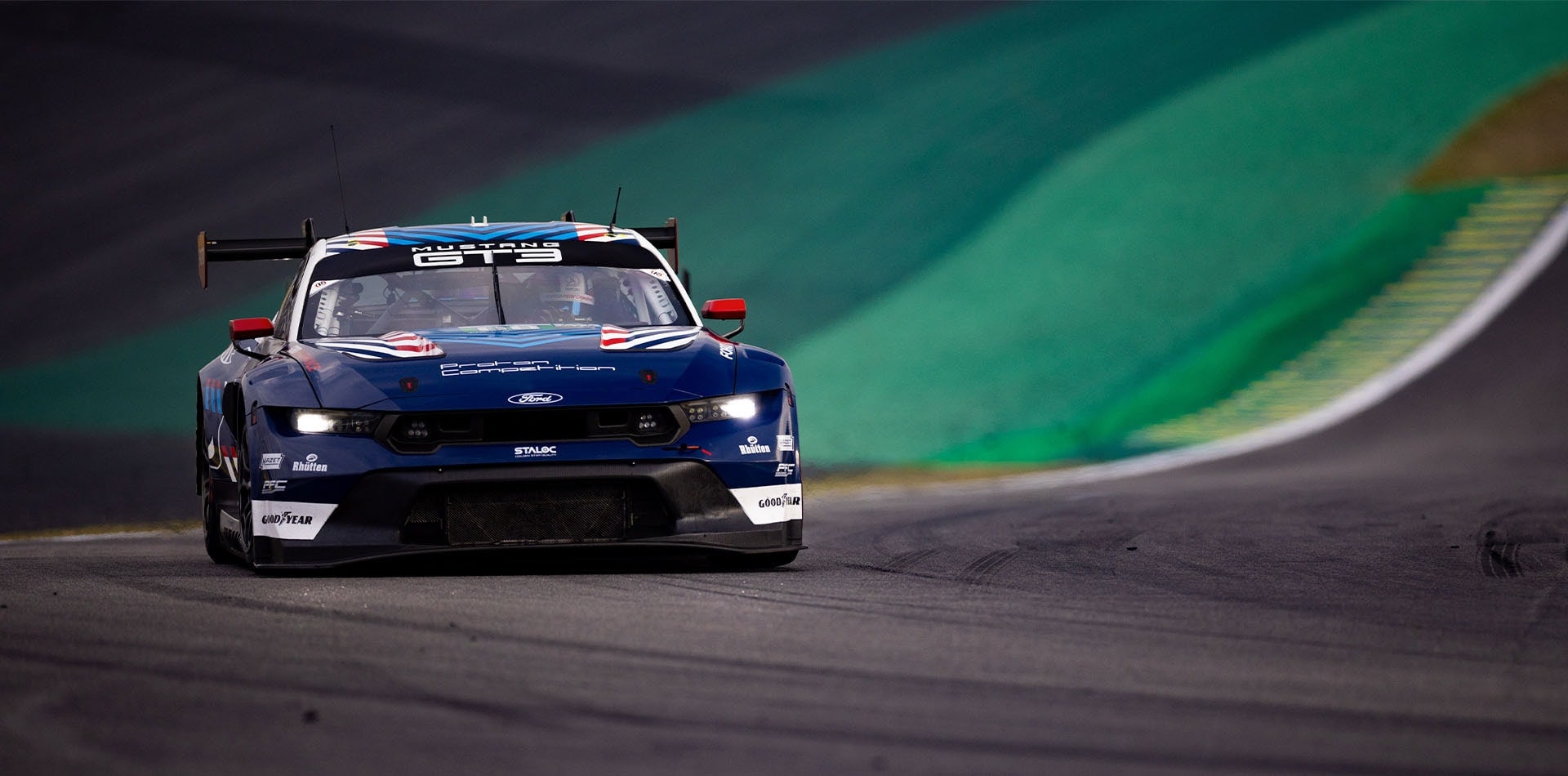 Spate vibrant al unui Ford Mustang GT3 de curse pe timp de noapte, cu stopurile roșii puternic iluminate și logo-ul Ford vizibil pe aripa spate, sugerând o atmosferă dinamică de cursă la Le Mans.